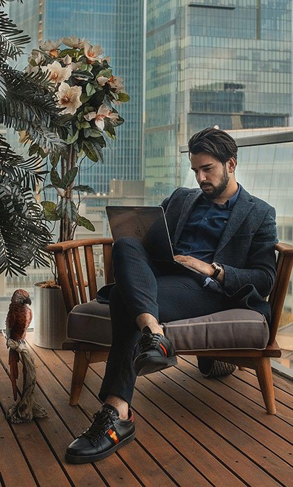 A Caucasian man sits in a chair, typing on a laptop. He's dressed in a dark blue jacket, blue pants, and a navy blue shirt.